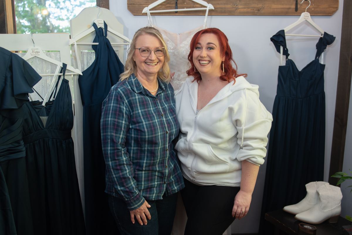Two women smile joyfully for the camera inside a boutique, surrounded by hanging dark blue dresses and a white wedding gown.