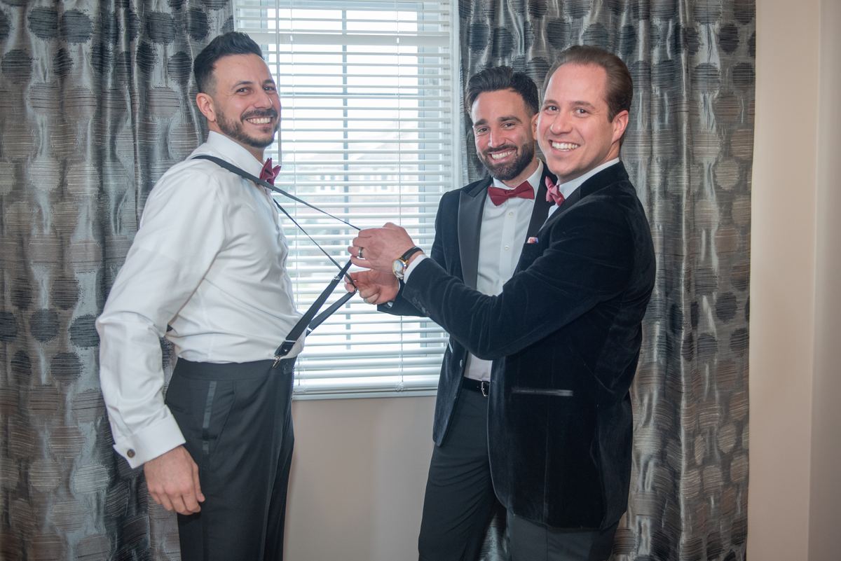Three formally dressed men smiling together as one adjusts another's suspenders in a warmly lit room.