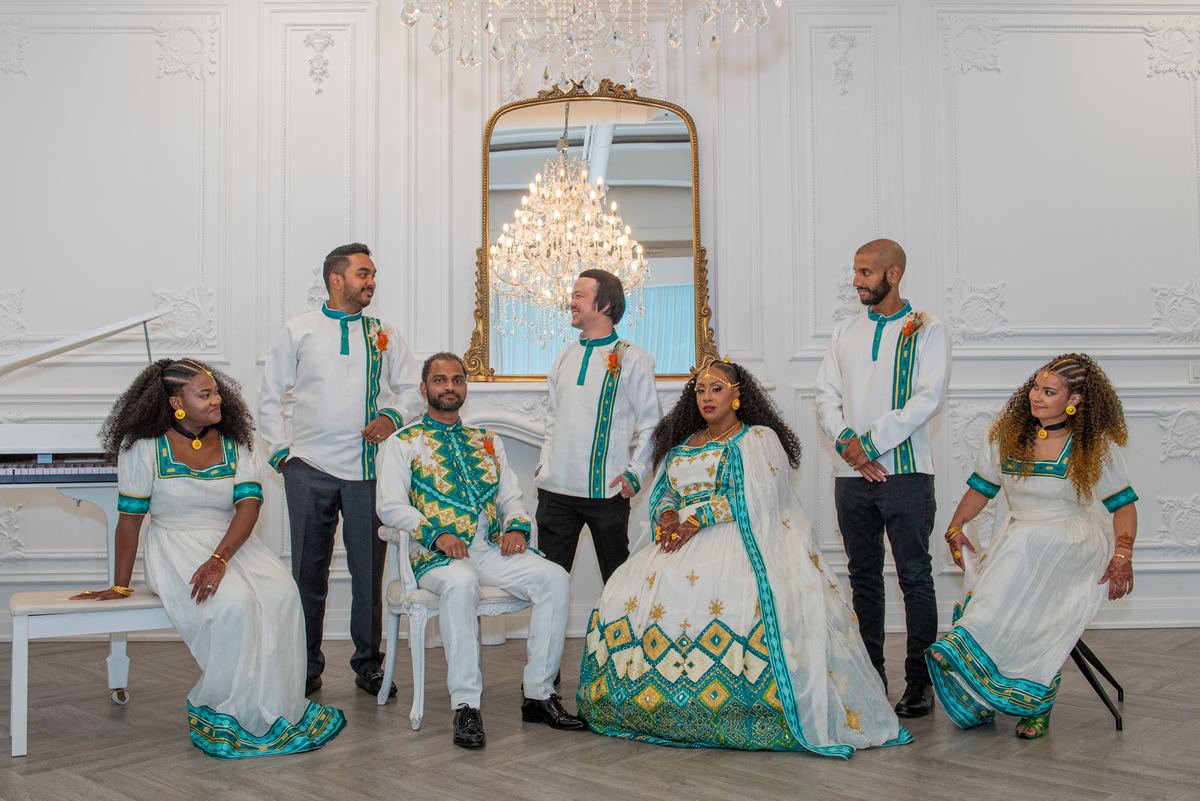 Seven people in white and teal traditional attire pose formally in an ornate room with a chandelier.