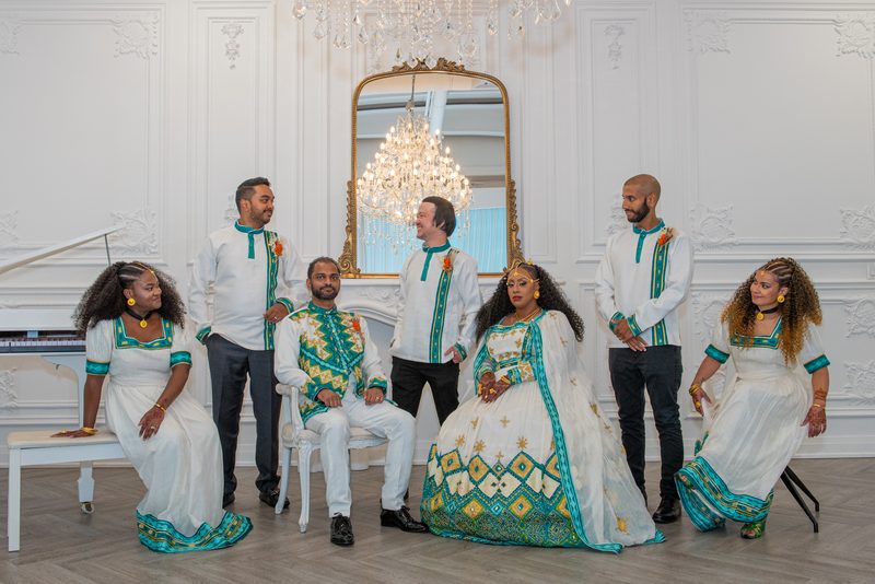 Seven people in white and teal traditional attire pose formally in an ornate room with a chandelier.