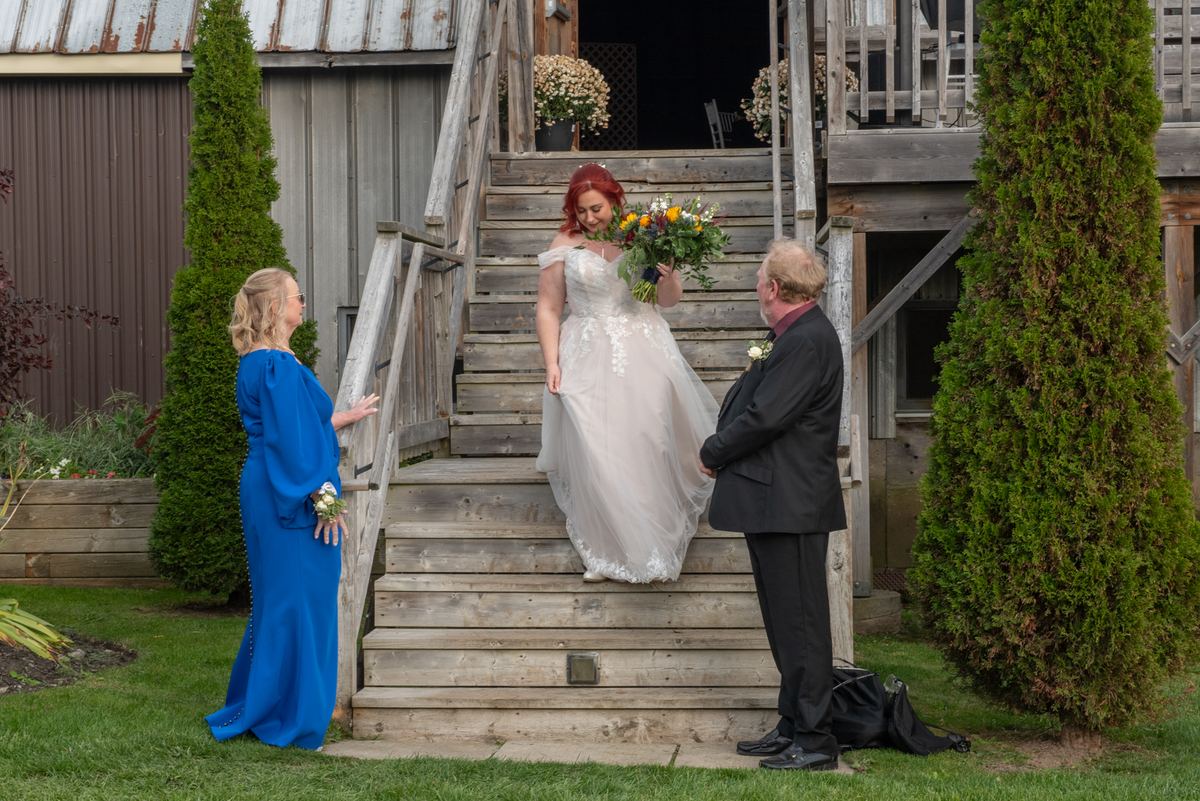 A red-haired bride descends wooden stairs, greeted by a woman in blue and a man in dark suit, as greenery frames the scene.