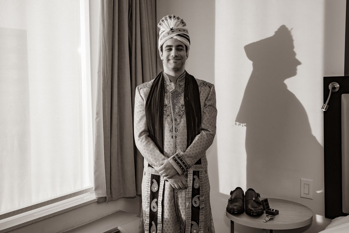 A groom in traditional Indian wedding attire stands smiling, his shadow cast dramatically against the wall beside him.