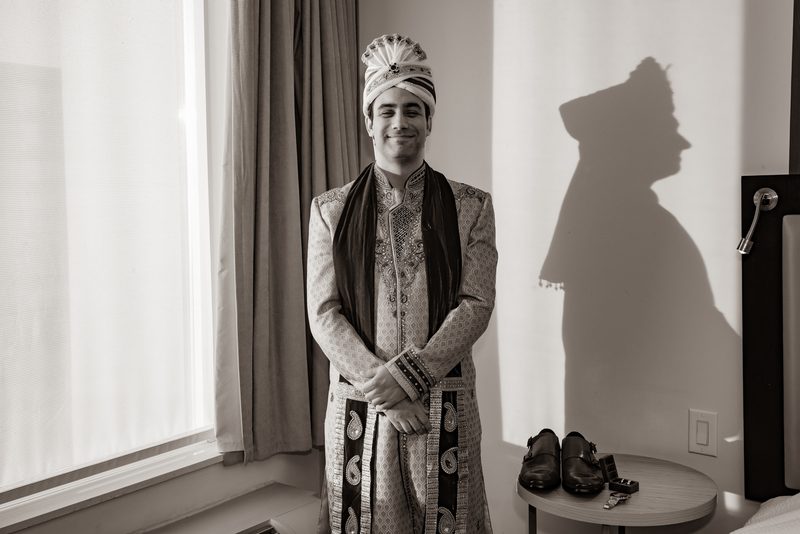 A groom in traditional Indian wedding attire stands smiling, his shadow cast dramatically against the wall beside him.