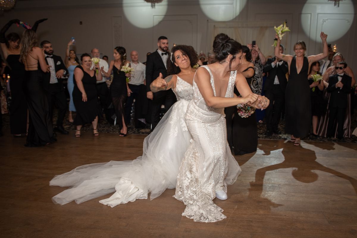 Two brides dance intimately on a wooden floor under spotlights, surrounded by cheering guests with phones.