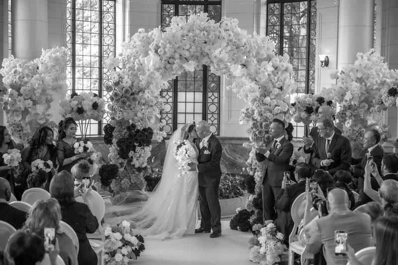 A bride and groom kiss at a wedding altar under a flower-draped arch, illuminated by soft light from tall windows.