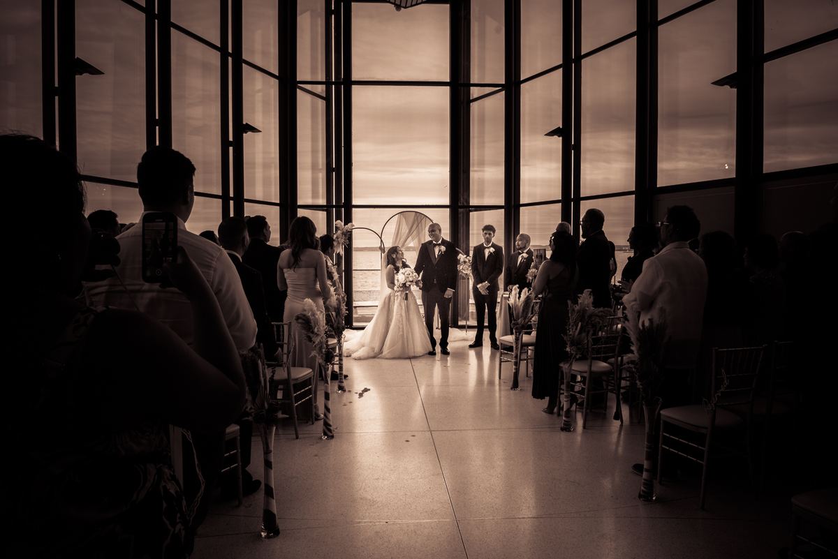 Bride and groom stand at the altar with a sunset over water visible through a glass wall, guests seated behind them.