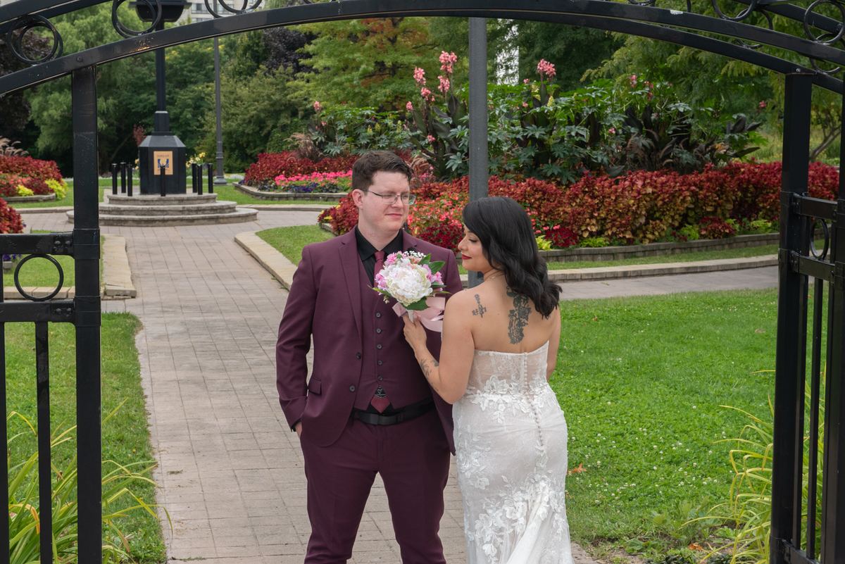 Bride in white lace gown and groom in burgundy suit share tender moment under ornate gate in garden.