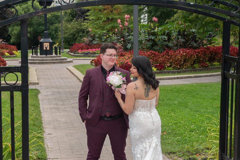 Bride in white lace gown and groom in burgundy suit share tender moment under ornate gate in garden.