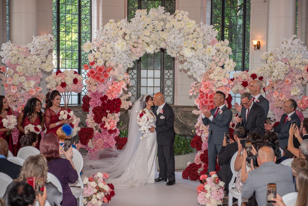 A bride and groom kiss under a floral arch, surrounded by guests taking photos in an ornate ceremony space.