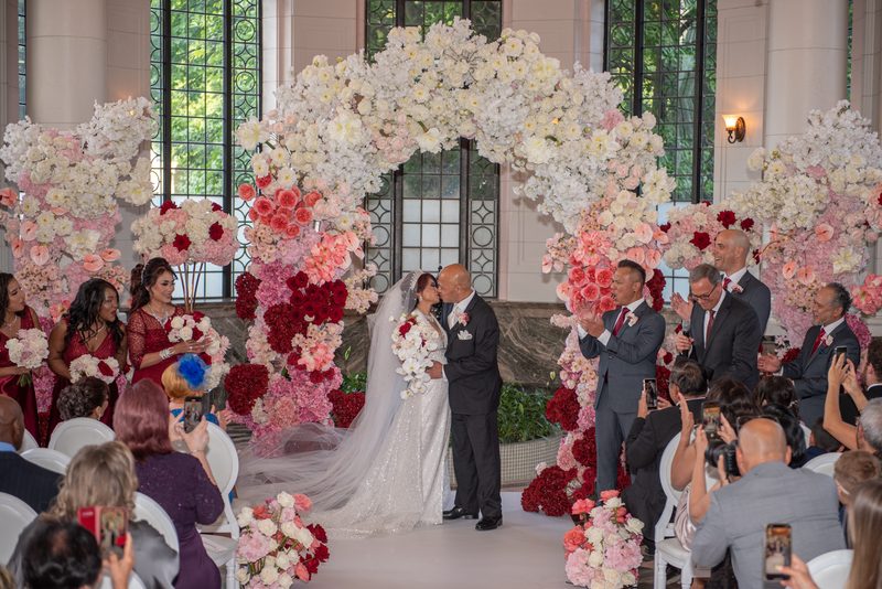 A bride and groom kiss under a floral arch, surrounded by guests taking photos in an ornate ceremony space.