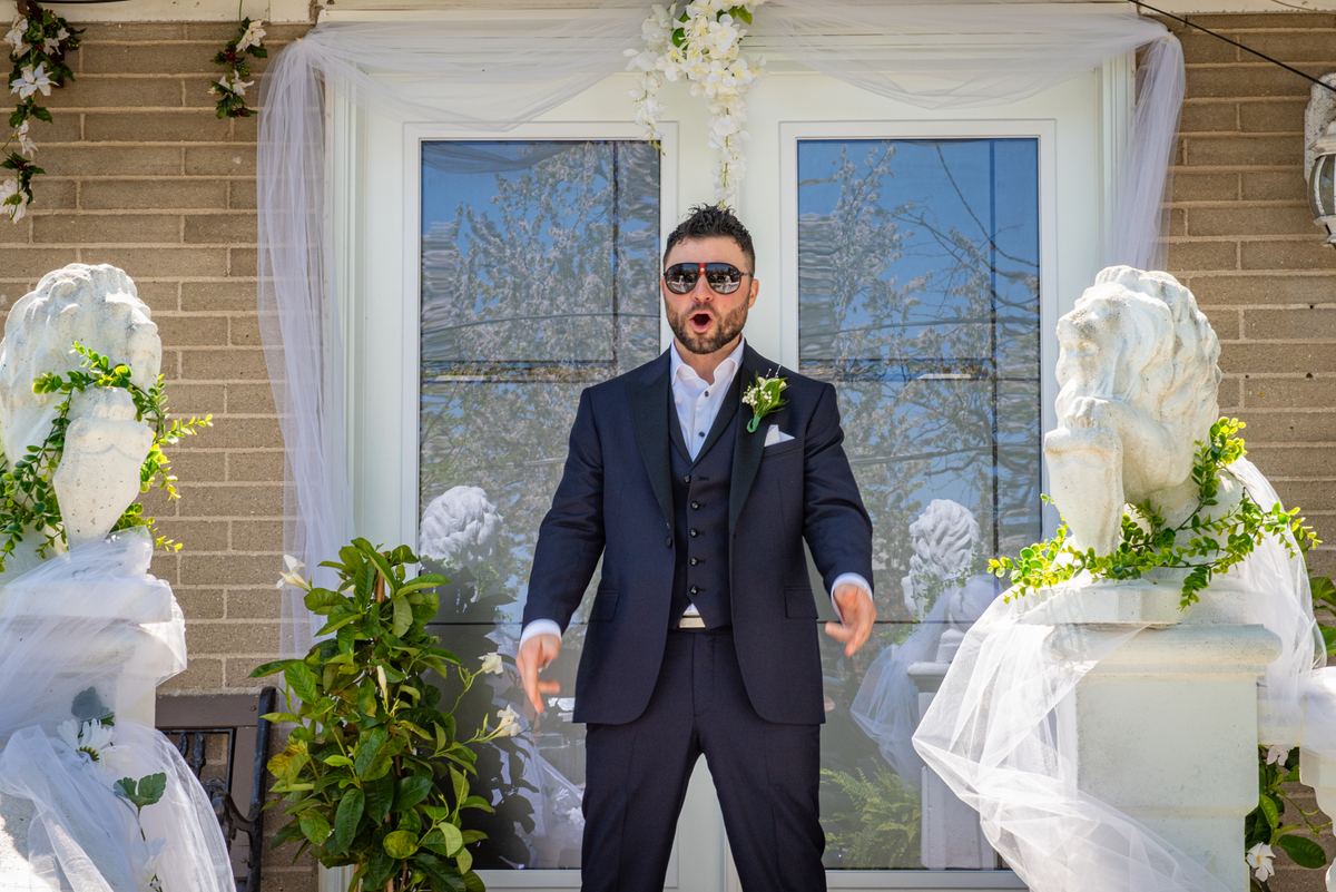 A surprised groom in a navy suit gestures outward at an outdoor wedding setup with white drapery and floral arrangements.