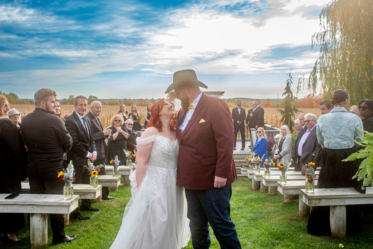 A bride and groom exchange vows outdoors, surrounded by guests on rustic benches in the late afternoon sun.
