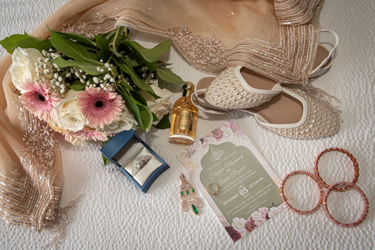 A bridal collection featuring a beaded gown, pearl shoes, flowers, ring, perfume, and invitation on a table.