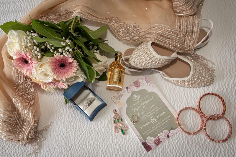 A bridal collection featuring a beaded gown, pearl shoes, flowers, ring, perfume, and invitation on a table.