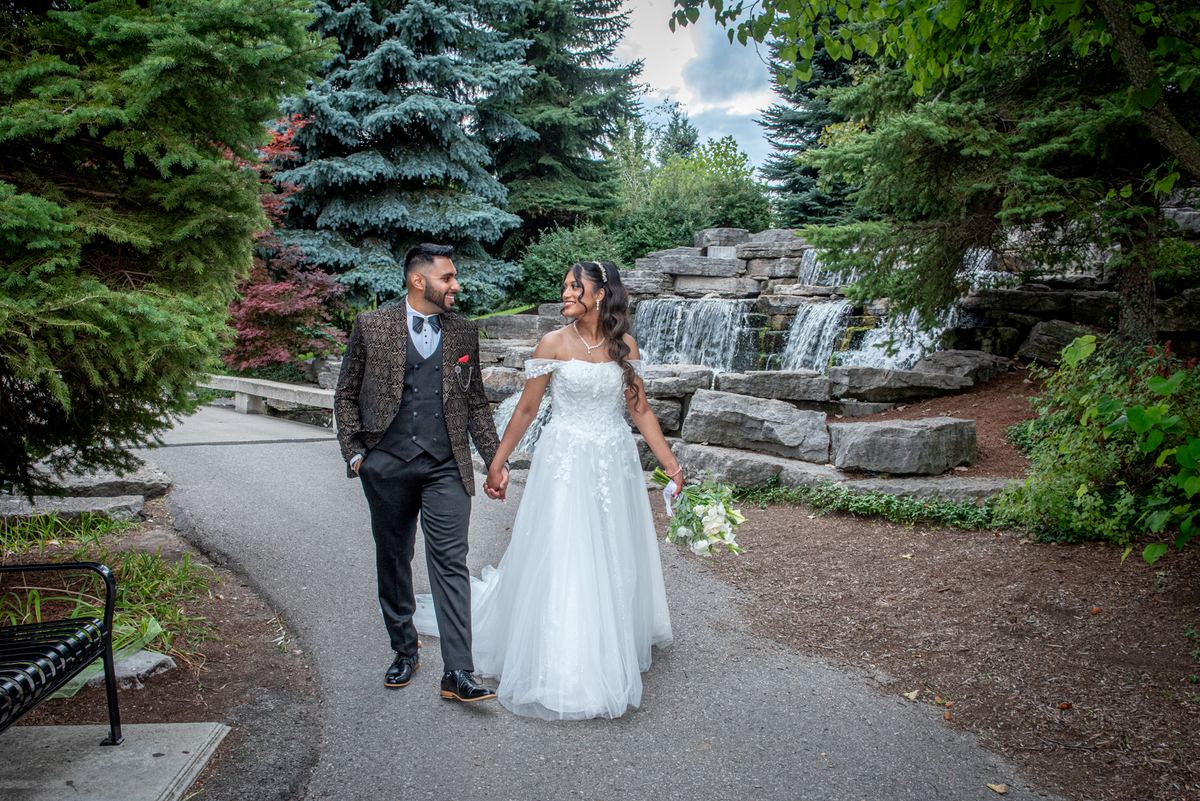 A newlywed couple walks hand-in-hand along a park path, smiling past a stone waterfall and autumn foliage.