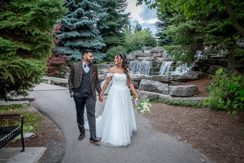 A newlywed couple walks hand-in-hand along a park path, smiling past a stone waterfall and autumn foliage.