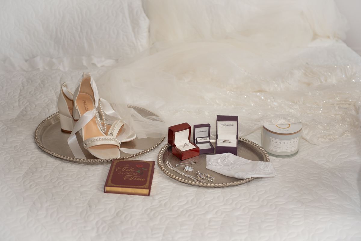 Wedding shoes, rings in boxes, lace glove, and candle arranged on trays next to a white quilted bed.