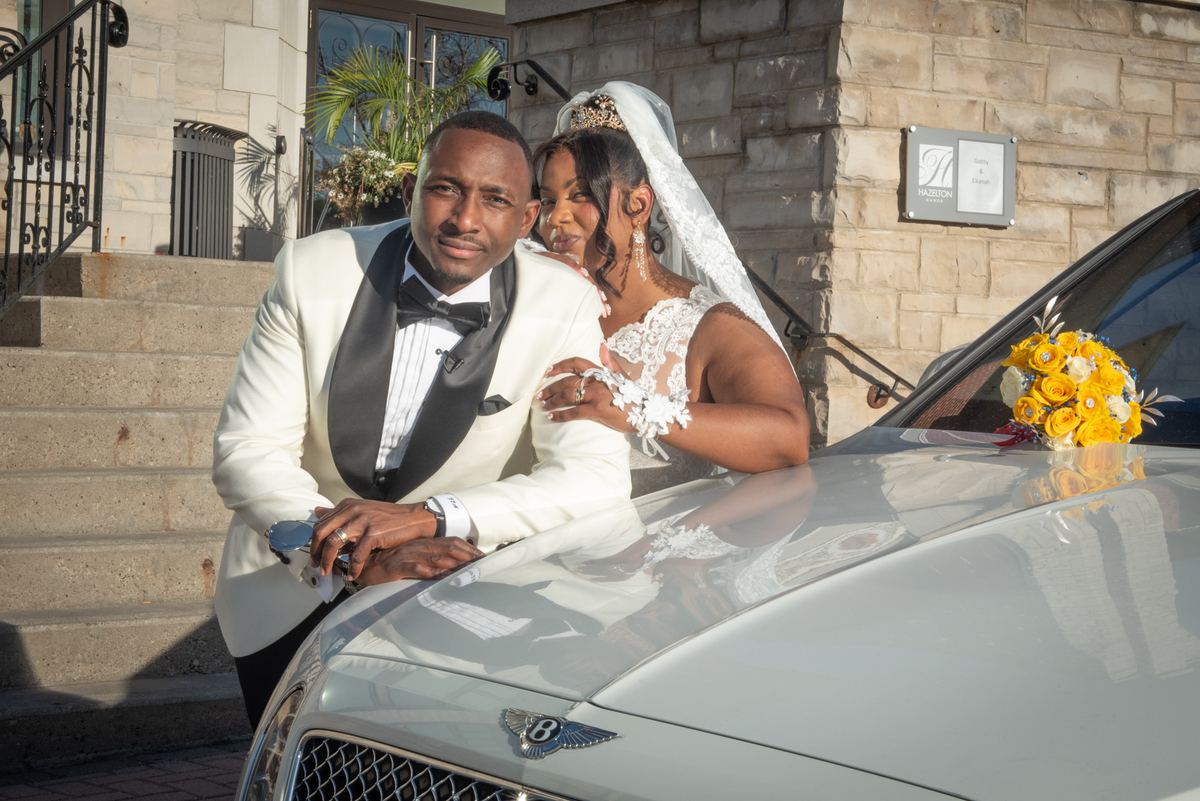 A bride in a lace gown and veil, with a yellow bouquet, and her groom in a white tuxedo stand beside a luxury Bentley.