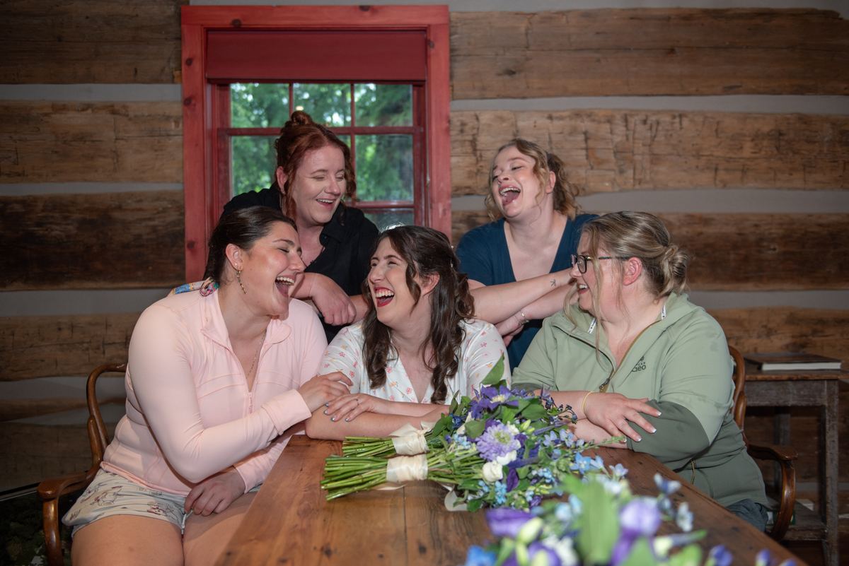 Five women laugh together around a wooden table in a rustic cabin, illuminated by natural light through a red-framed window.