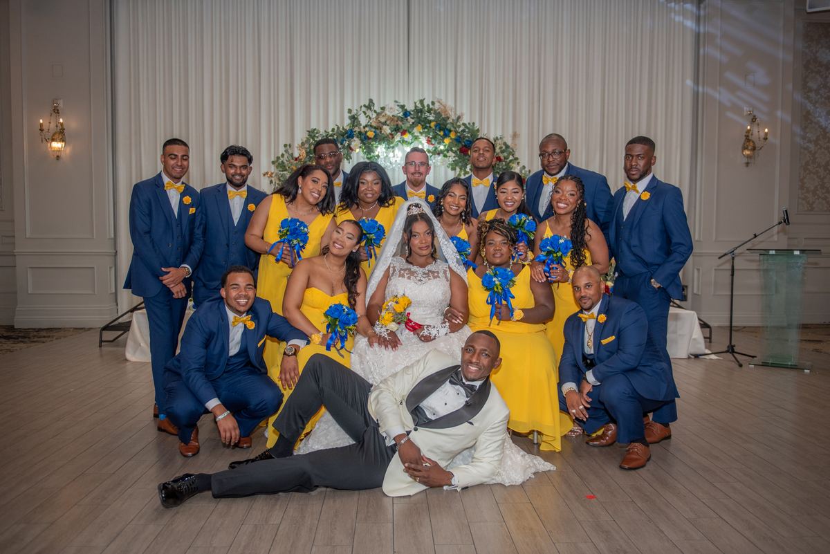 A bride and groom smile at the centre of their wedding party, bridesmaids in yellow and groomsmen in navy, in a ballroom.