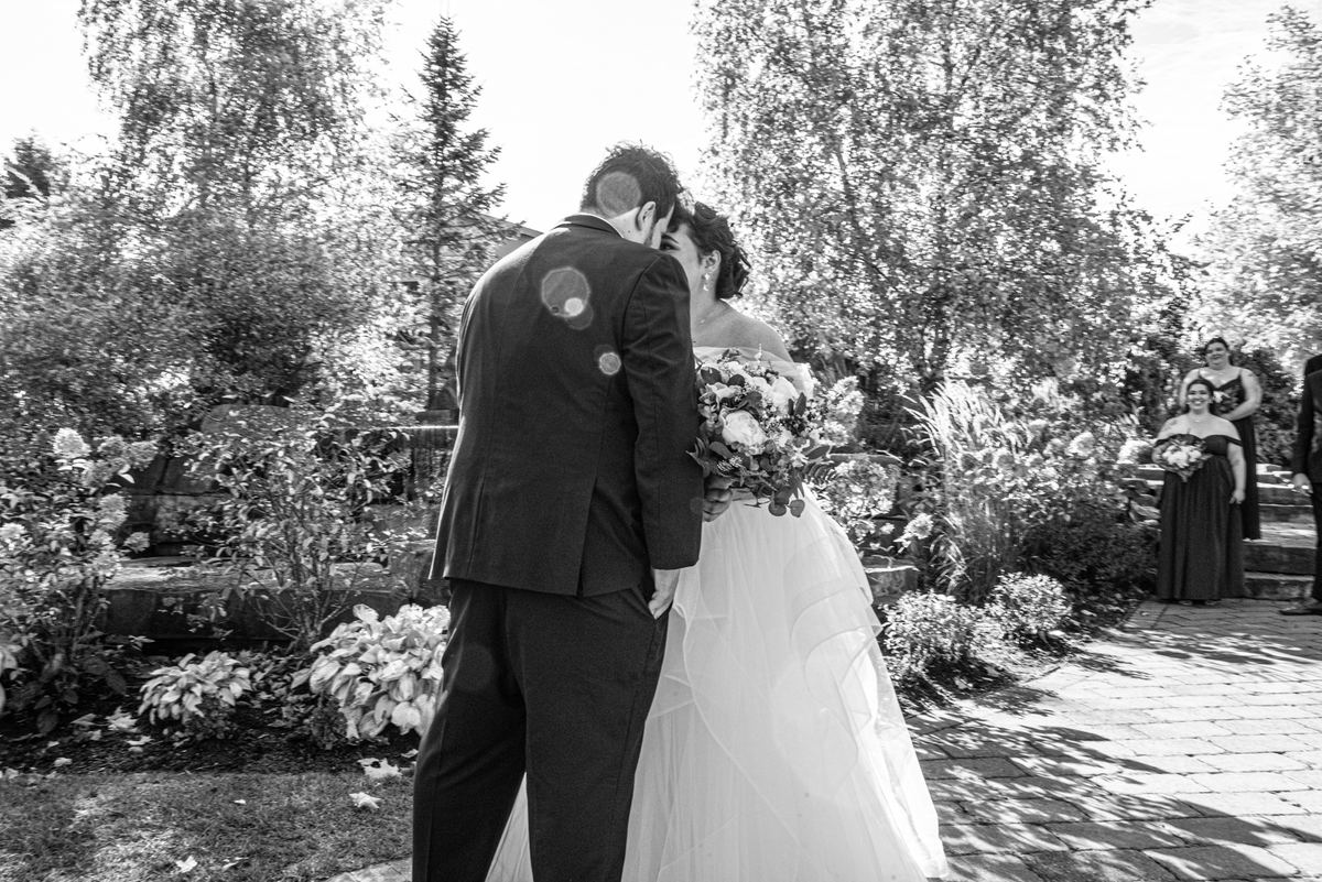 A bride and groom kiss in a sunlit garden with bridesmaids observing from behind, creating lens flare.