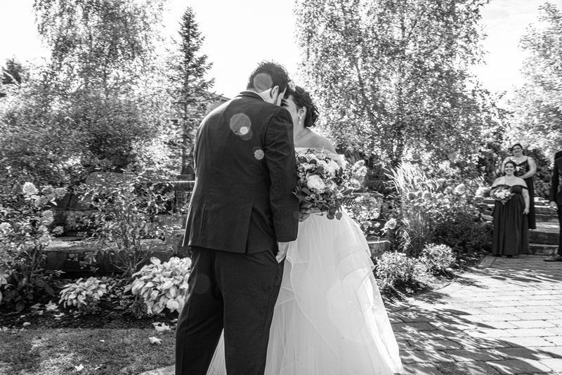 A bride and groom kiss in a sunlit garden with bridesmaids observing from behind, creating lens flare.