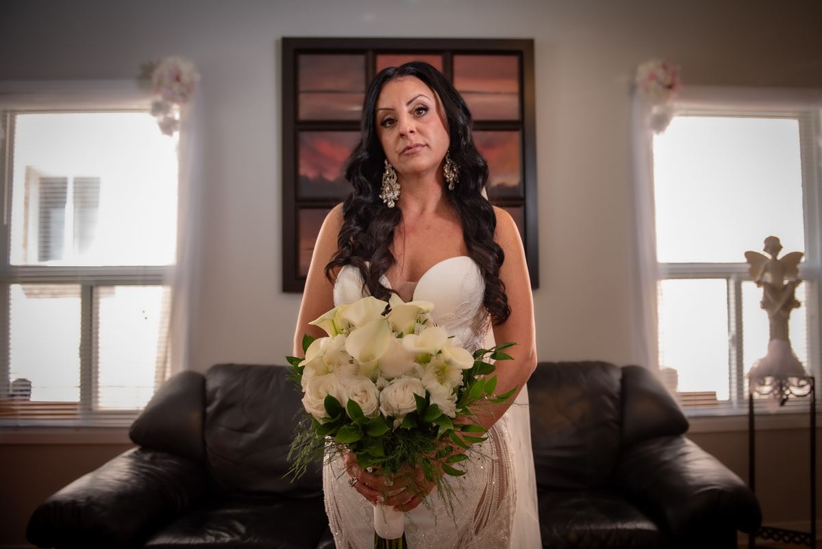 A bride in a wedding gown holds white flowers, standing thoughtfully near a window-lit room with decor elements.