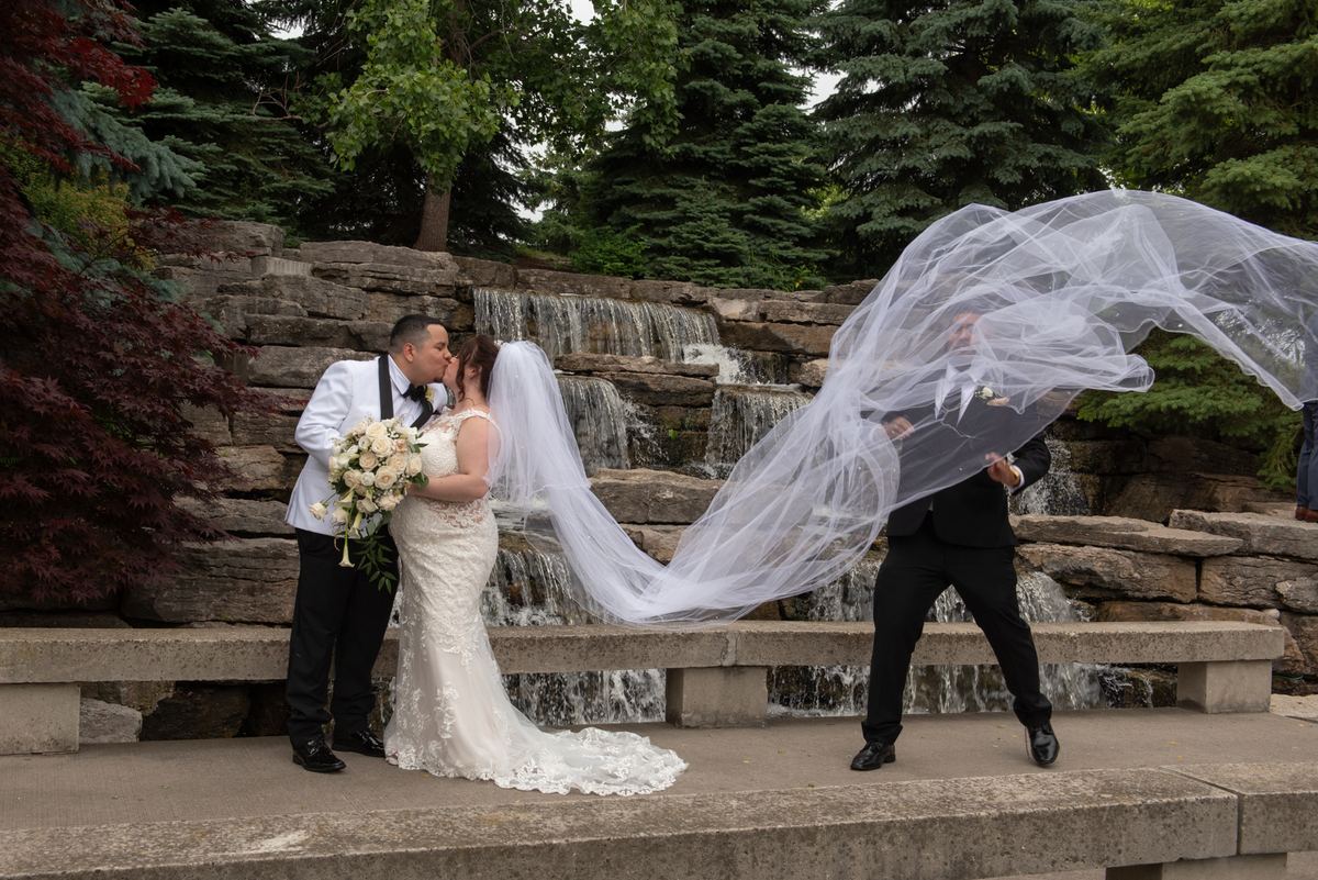 Bride kisses groom beside stone waterfall, holding back billowing veil amid lush greenery and layered steps.