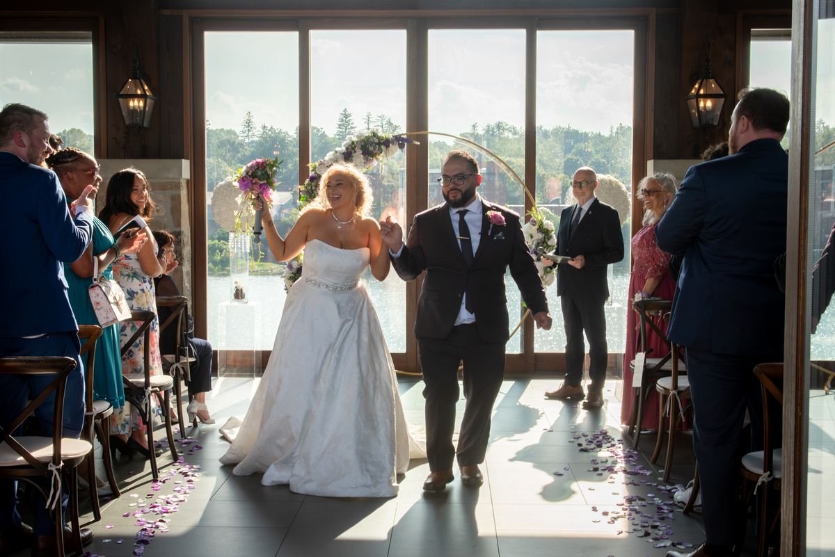 Bride and groom walk down an aisle lined with purple petals, framed by an arched backdrop, as guests watch.
