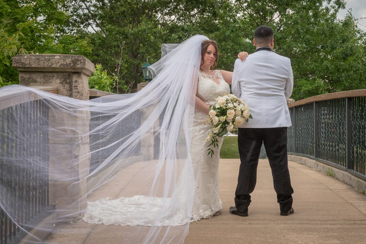Bride in white gown and veil stands with groom on stone bridge, arm around her shoulder, against green tree backdrop.