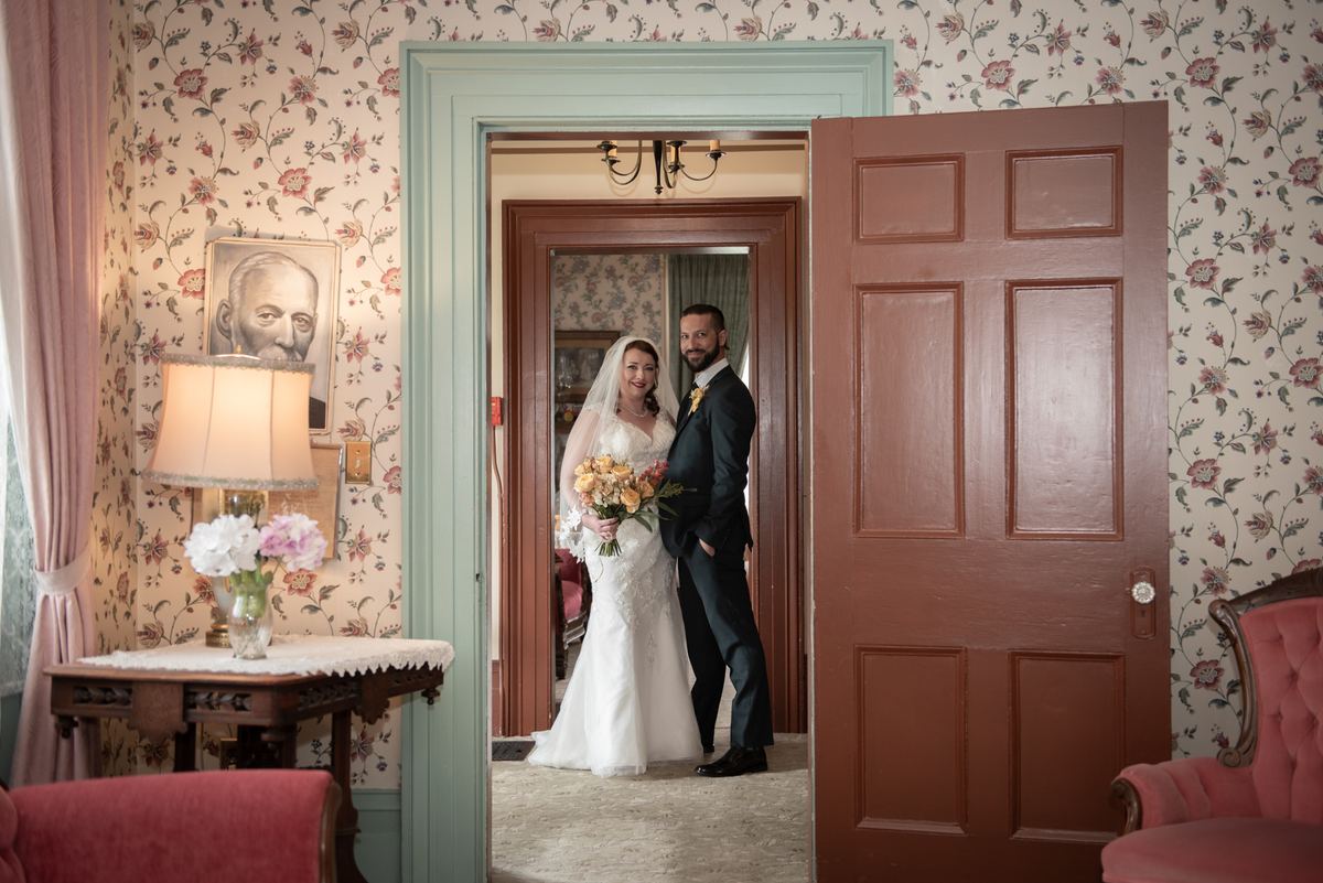 A bride and groom stand in an open doorway, smiling warmly against vintage floral wallpaper and woodwork.