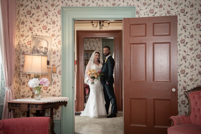 A bride and groom stand in an open doorway, smiling warmly against vintage floral wallpaper and woodwork.