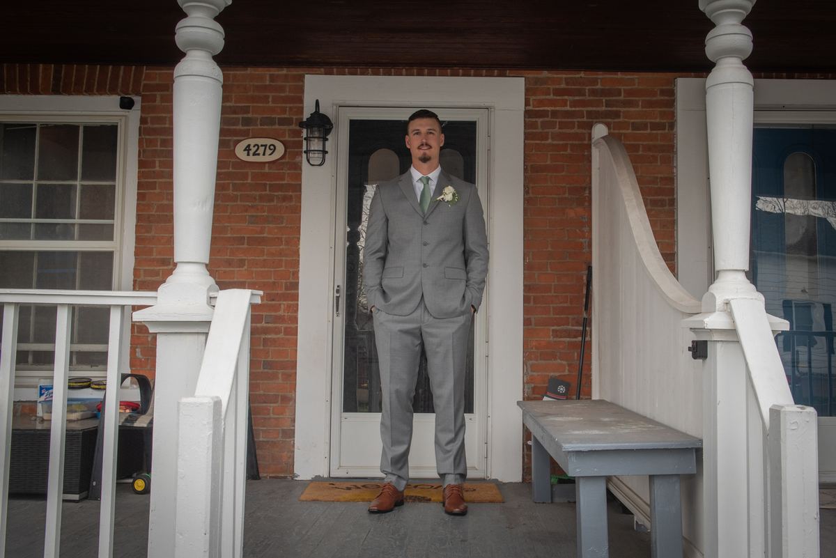 A groom in a gray suit with boutonnière stands on the porch of a brick house, smiling at the camera.