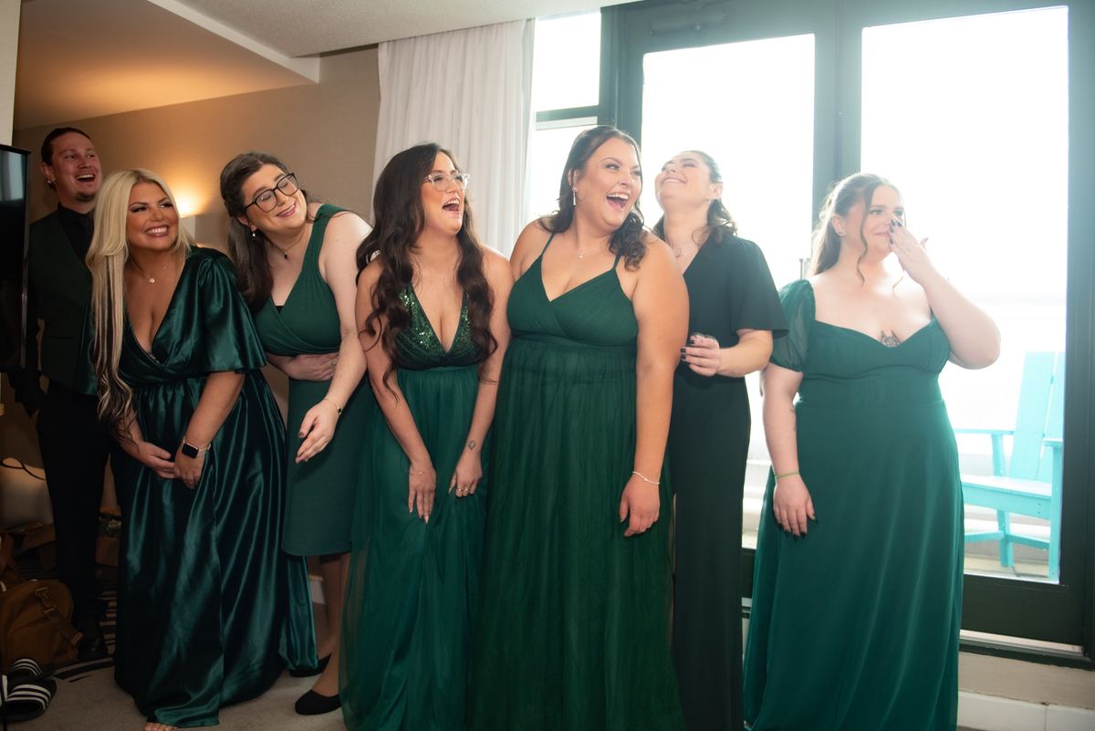 Bridesmaids and one groomsmen laugh together in bright light, dressed in emerald green gowns and dark suit.