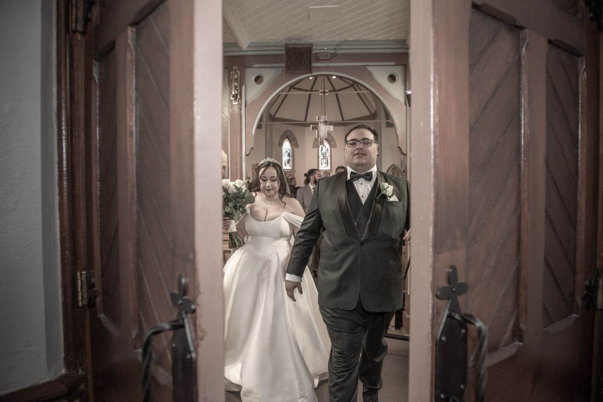 A bride with a bouquet and a groom in a tuxedo walk down a church aisle framed by open wooden doors and soft window light.