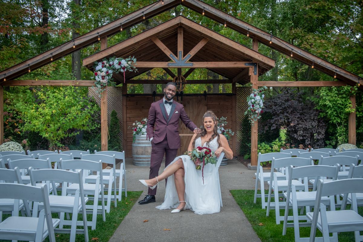 A smiling groom stands beside a laughing bride seated under a rustic wooden arbor adorned with flowers.