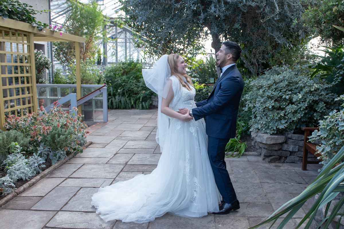 A smiling bride and groom laugh together in a greenhouse garden, their hands clasped, surrounded by lush greenery.