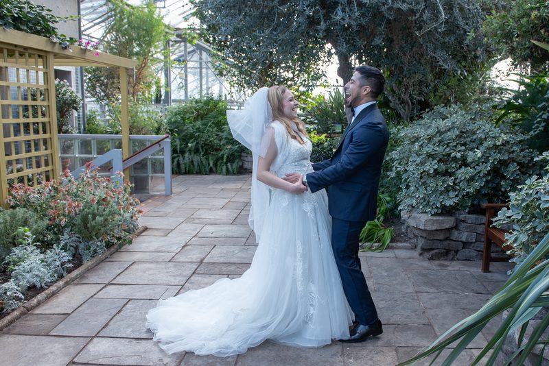 A smiling bride and groom laugh together in a greenhouse garden, their hands clasped, surrounded by lush greenery.