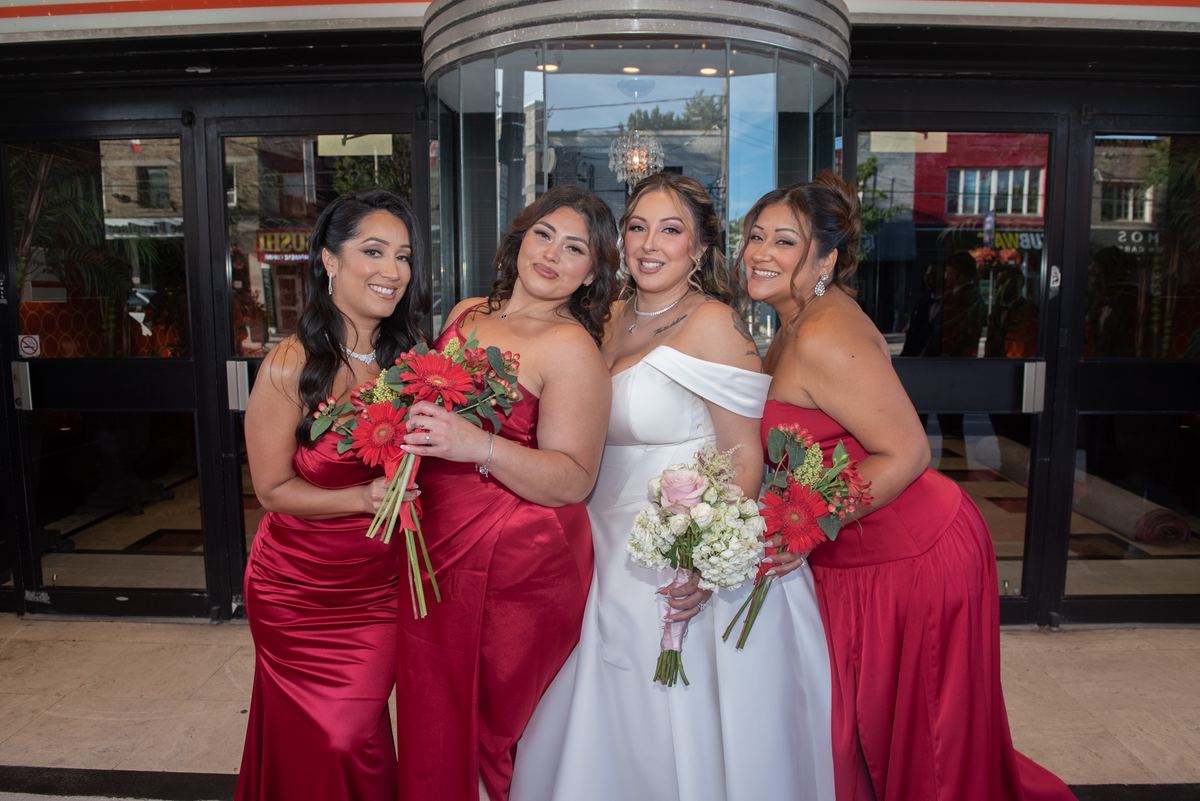 Four women, including a bride in a white off-the-shoulder gown, pose with red and white flower bouquets outside a building.