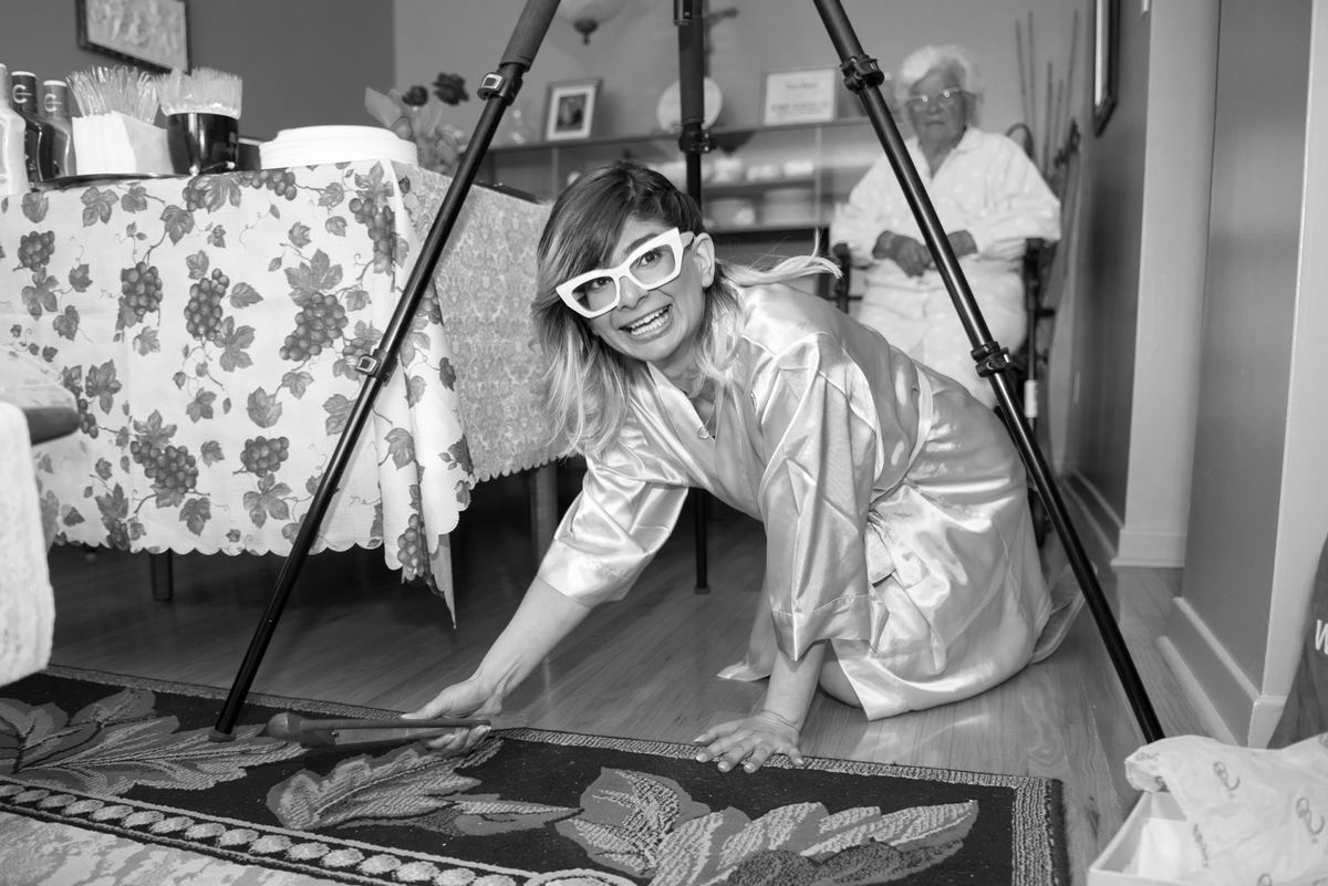 A woman in a satin robe holds a broomstick and crawls playfully while smiling at the camera during a staged photo shoot.