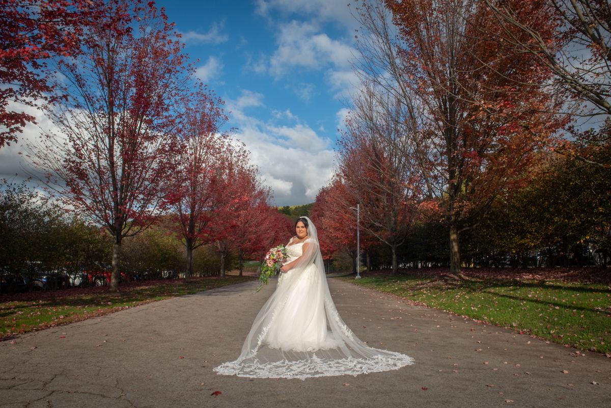 A bride in a white gown and veil stands on a path surrounded by autumn trees, holding a bouquet and gazing at the camera.