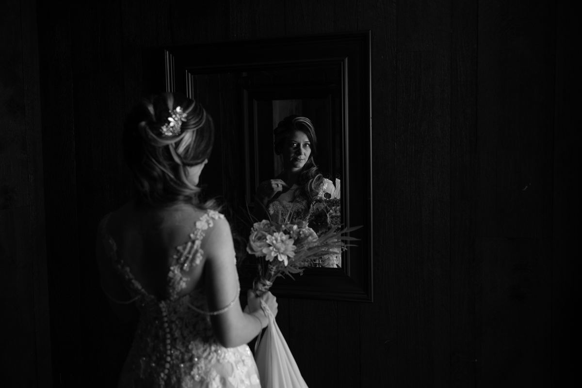 A bride stands before a mirror in a dimly lit room, holding her bouquet and gazing at her reflection.