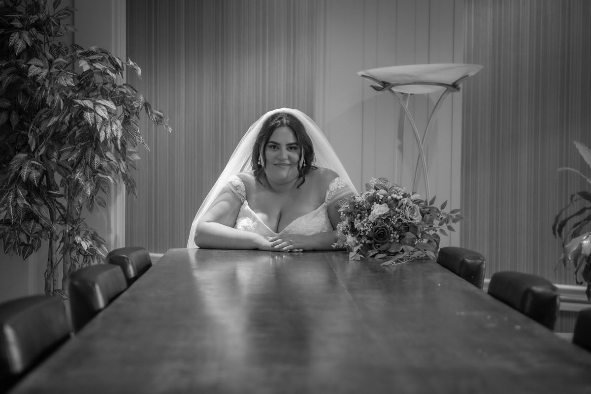 A smiling bride rests her arms on a wooden table in a modern room with potted plants and ambient lighting, bouquet.