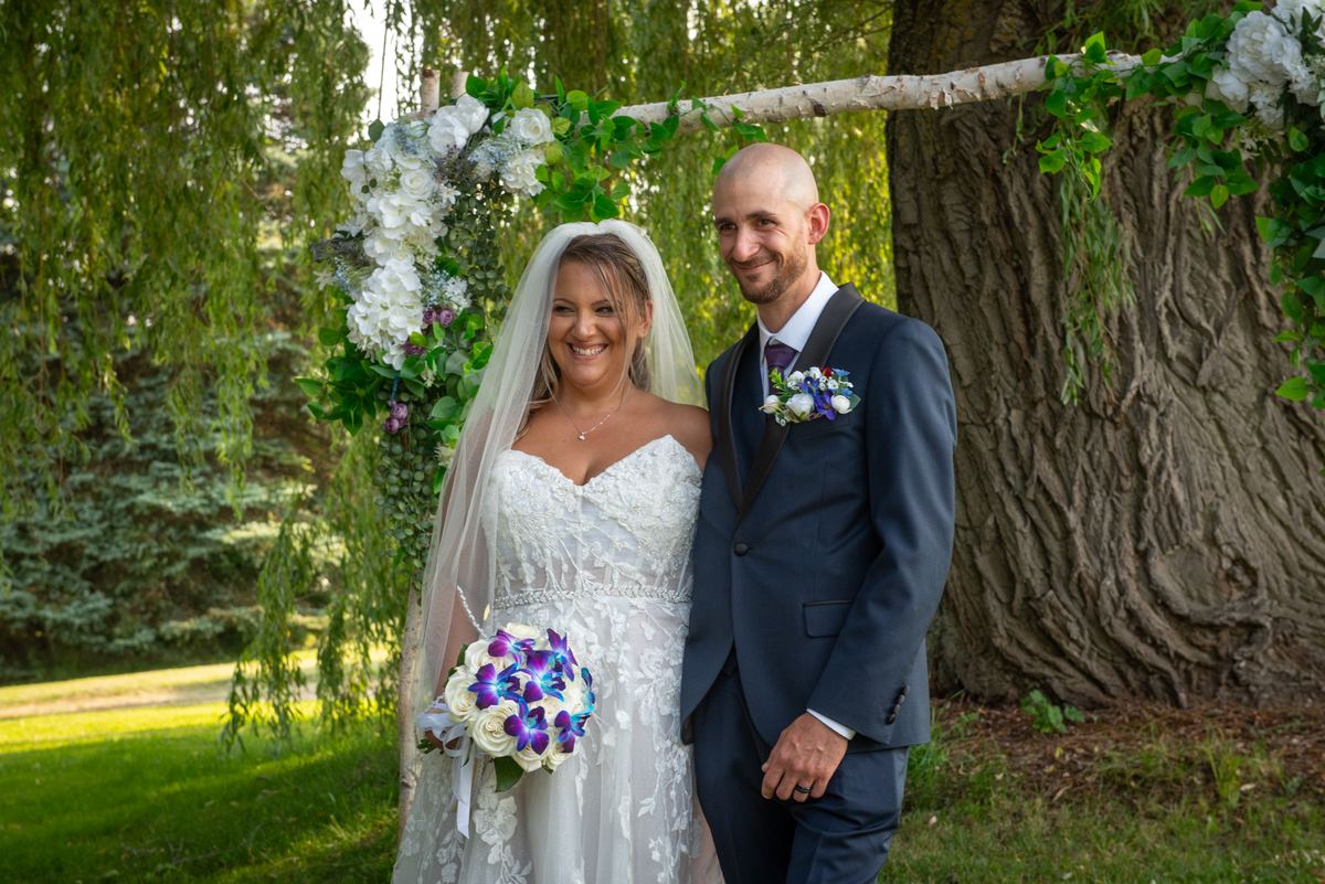 A smiling bride and groom stand beneath a floral arch in dappled sunlight, with the grassy ground casting gentle shadows.