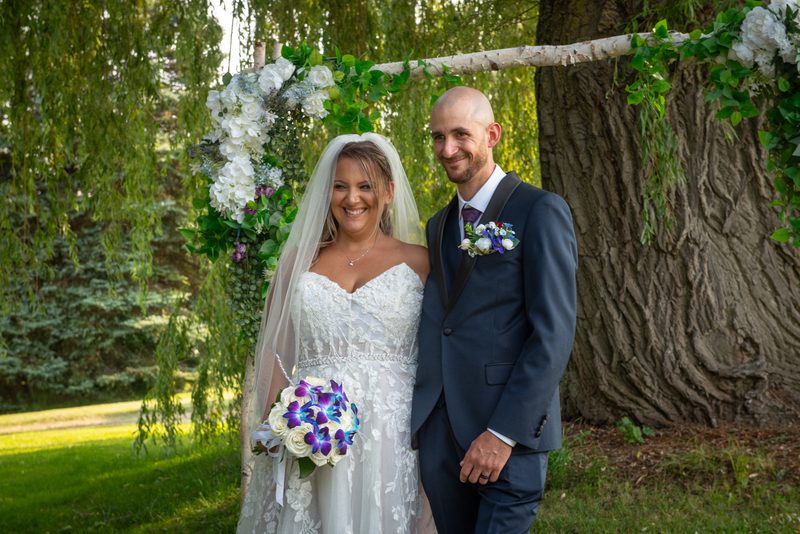 A smiling bride and groom stand beneath a floral arch in dappled sunlight, with the grassy ground casting gentle shadows.