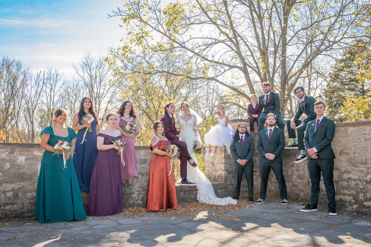 A wedding party poses outdoors, surrounded by autumn trees and warm light, in various outfits including a white gown.