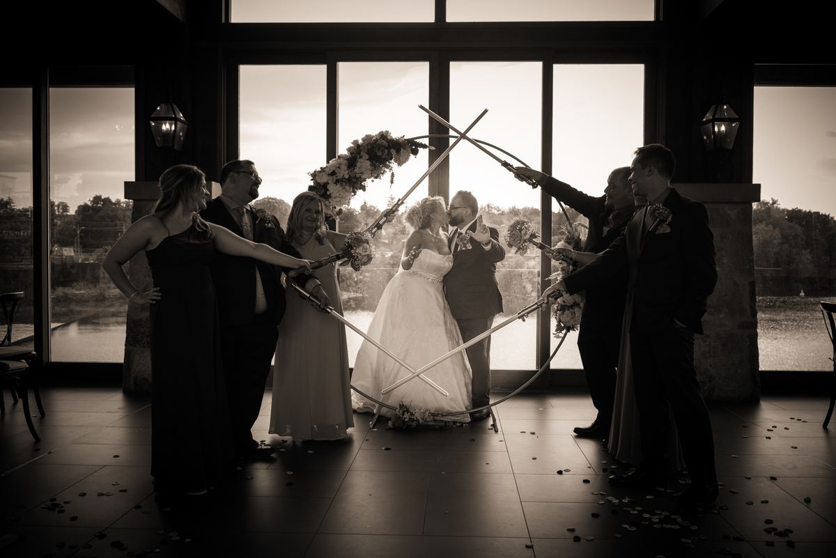A bride and groom kiss as wedding party members wield curved swords in a playful dance, surrounded by petals.