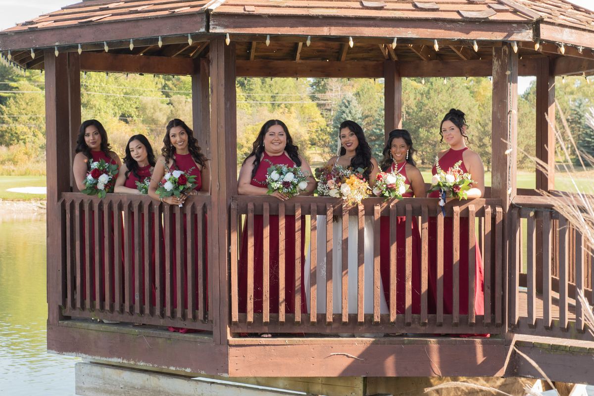 A bride and her bridesmaids stand in a wooden gazebo by water, holding flowers, bathed in golden light.