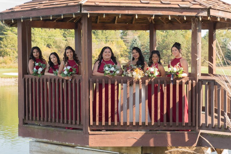 A bride and her bridesmaids stand in a wooden gazebo by water, holding flowers, bathed in golden light.