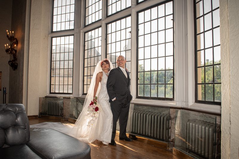 A bride in white and a groom in dark suit stand near windows, bathed in golden light, smiling at the camera.